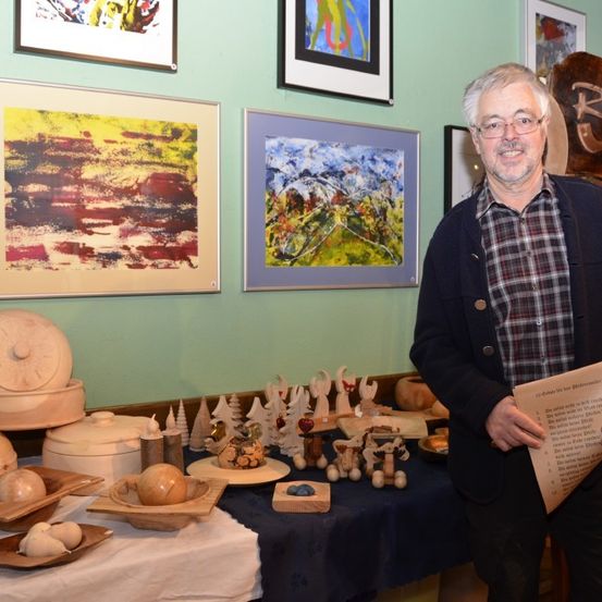 Ein älterer Mann mit Brille steht vor einem Tisch voller verschiedener Holzobjekte. Hinter ihm hängen mehrere gerahmte Gemälde an der Wand. Er hält ein Stück Papier in der Hand.