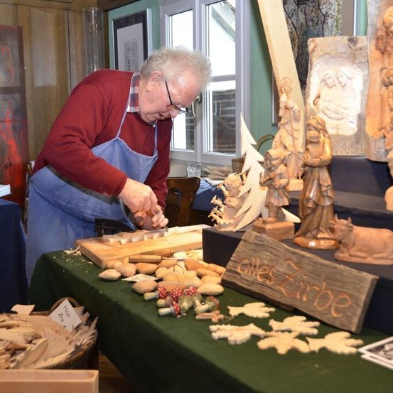 Ein älterer Mann mit Brille und Schürze schnitzelt ein Holzobjekt auf einem Tisch mit verschiedenen Holzgegenständen. Hinter ihm steht eine Holzskulptur von Jesus Christus und eine Holzskulptur einer Kuh.