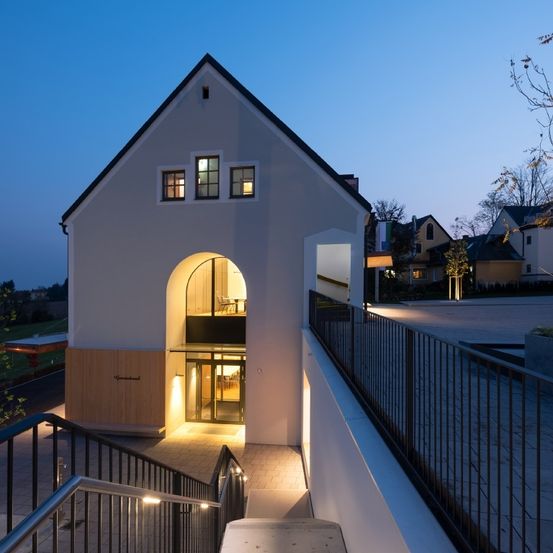 Ein modernes Haus mit einem gewölbten Eingang, beleuchtet in der Nacht, mit einem Balkon mit Tisch und Stühlen, umgeben von einem Metallzaun.
