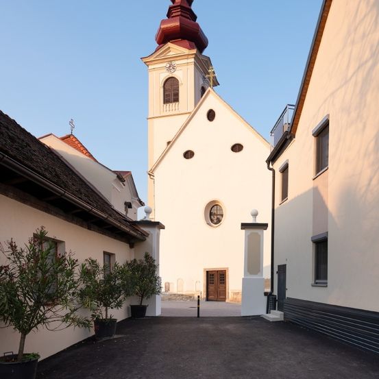 Eine Kirche mit rotem Dach und Turm steht vor zwei Gebäuden. Die Kirche hat eine Uhr und ein Kreuz auf der Spitze.