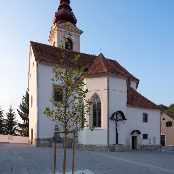 Eine weiße Kirche mit einem braunen Dach, einem Turm mit einem Spitzturm und einem Baum davor. Ein Kruzifix ist an der Wand angebracht.