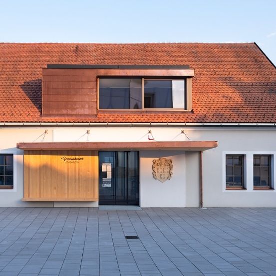 Ein modernes Gebäude mit einem roten Ziegeldach und einer weißen Außenfassade, das eine kupferfarbene Überdachung über dem Eingang aufweist.