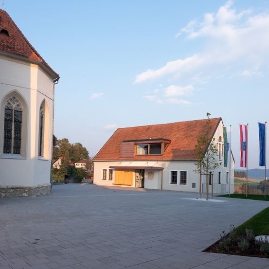 Ein leerer Innenhof vor einer Kirche mit einer gelben Tür. Der Himmel ist blau mit einigen Wolken. Im Vordergrund befinden sich drei Flaggen und ein Baum.