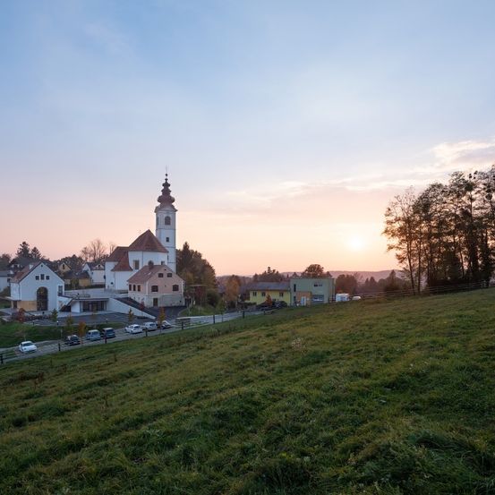 Eine malerische Ansicht eines Dorfes mit einer Kirche, Häusern und geparkten Autos, umgeben von Bäumen und Gras unter einem blauen Himmel mit Wolken.