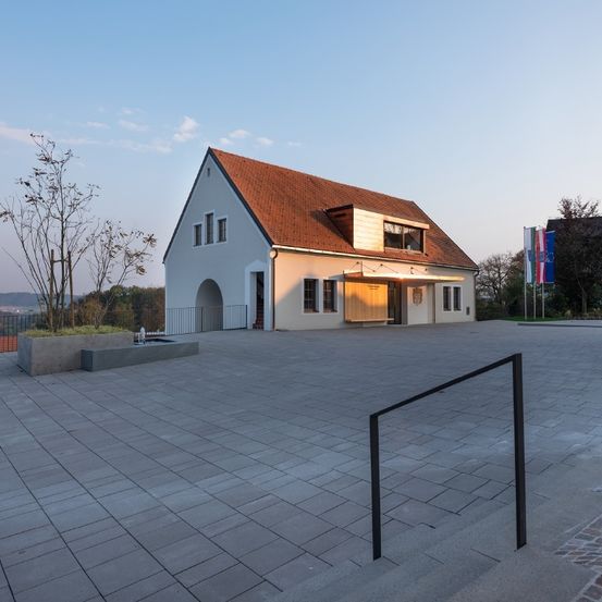 Ein weißes Gebäude mit rotem Dach und einem gewölbten Eingang. Es ist von einem gepflasterten Bereich mit einem Metallgeländer umgeben. Es gibt einen Baum in einem Betonpflanzer und Flaggen im Hintergrund.