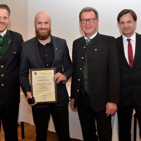 Vier Männer in Anzügen stehen hinter einem Tisch, einer hält ein Zertifikat mit 'Unkunde' darauf, lächeln für ein Foto.