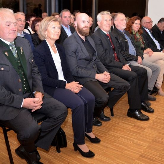 Mehrere Personen sitzen in einem Saal, leger gekleidet. Sie scheinen an einer Veranstaltung teilzunehmen. Die Person in der Mitte hat einen Bart und trägt einen Anzug. Der Raum hat Holzböden.