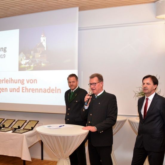 Eine formelle Zeremonie mit drei Männern in Anzügen. Ein Mann hält ein Mikrofon und spricht, während ein anderer ihn ansieht. Eine Projektionsleinwand zeigt ein Bild einer Kirche und das Datum 2019. Zertifikate liegen auf einem nahen Tisch.