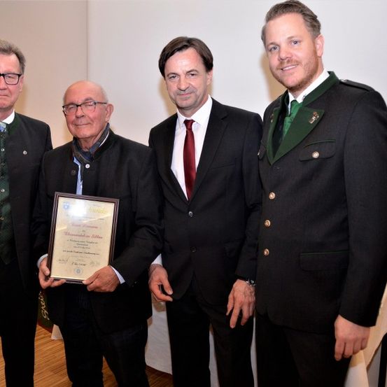 Vier Männer stehen vor einer weißen Wand und lächeln für ein Foto. Der Mann in der Mitte hält ein gerahmtes Zertifikat. Rechts neben einem Mann in einem roten Anzug steht ein Mann in einem grün-schwarzen Jackett. Links hält ein Mann mit einer Brille das Zertifikat.