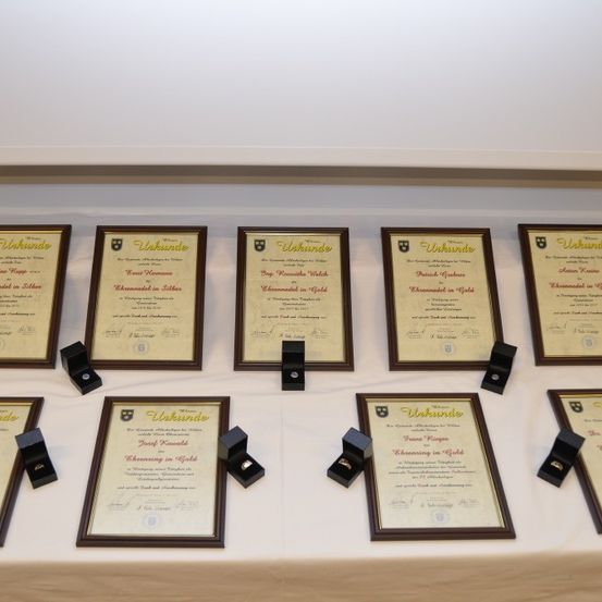 Mehrere gerahmte Urkunden und kleine schwarze Schmuckkästchen sind auf einer weißen Oberfläche ausgestellt. Die Urkunden sind goldfarben mit Text, und die Kästchen enthalten Ringe.