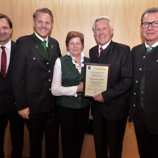 Fünf Personen stehen lächelnd für ein Foto zusammen. Die zentrale Figur ist eine Frau, die eine gerahmte Urkunde hält. Der Mann neben ihr hält den Rahmen der Urkunde.