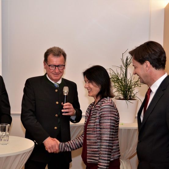 Ein Mann mit einem Mikrofon spricht zu einer lächelnden Frau in einer gestreiften Jacke, während zwei andere Männer in der Nähe stehen. Sie befinden sich in einem Raum mit weißen Wänden und einem Tisch mit einer Topfpflanze.