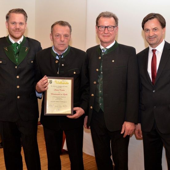 Vier Männer stehen in formeller Kleidung; einer hält eine gerahmte Urkunde. Sie lächeln und scheinen bei einer Preisverleihung zu sein. Der Mann in der Mitte hält die Urkunde, und der Mann rechts trägt eine rote Krawatte.