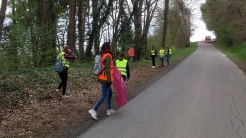 Eine Gruppe von Freiwilligen in Reflektorwesten und mit Müllsäcken geht an einer von Bäumen gesäumten Straße entlang. Ein rotes Fahrzeug ist in der Ferne sichtbar.