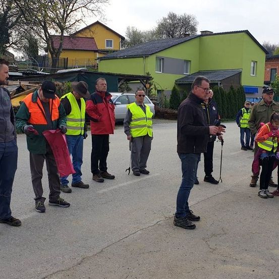 Mehrere Menschen stehen auf einer Straße, tragen Warnwesten, einer der Männer hält ein Mikrofon. In der Nähe ist ein Haus mit grüner Fassade zu sehen.