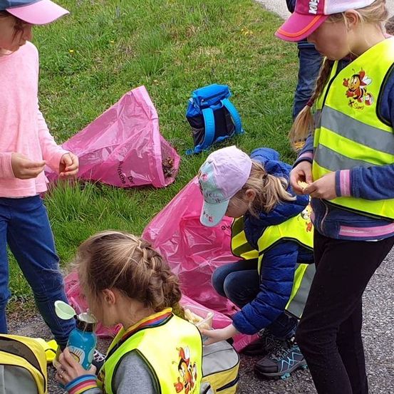 Kinder in Warnwesten versammeln sich im Freien, einige gebeugt und sammeln Müll, während andere in der Nähe stehen. Ein pinker Müllsack liegt auf dem Boden.