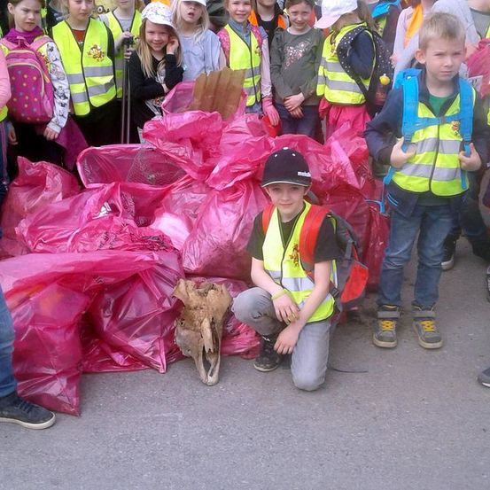 Eine Gruppe von Kindern in Sicherheitswesten steht um einen Haufen rosa Säcke herum, möglicherweise Müll sammelnd. Ein Junge in einem Hut kniet neben einem Schädel.
