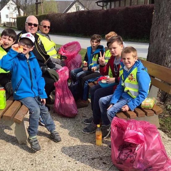 Eine Gruppe von Kindern und Erwachsenen sitzt auf einer Bank in einem Park. Sie tragen Warnwesten und haben Plastiktüten. Die Kinder essen Essen, und einer hält ein Friedenszeichen.