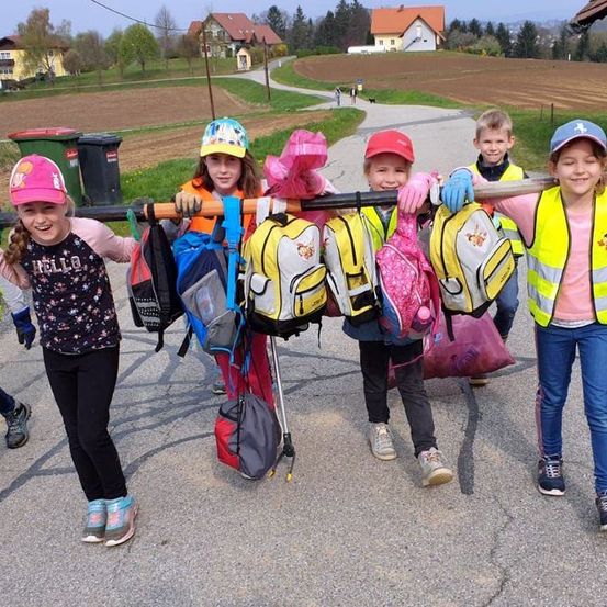 Eine Gruppe von Kindern, die Rucksäcke tragen, Hochsichtbarkeitswesten tragen und einen Stab mit Taschen halten. Sie stehen auf einem Weg mit Mülleimern, Häusern und einem Feld im Hintergrund.