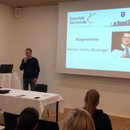 Ein Mann steht vor einer Projektionsleinwand, auf der eine Folie von Michael Fuchs-Wurzinger, dem Bürgermeister, zu sehen ist. Menschen sitzen um einen Tisch in einem Besprechungsraum.