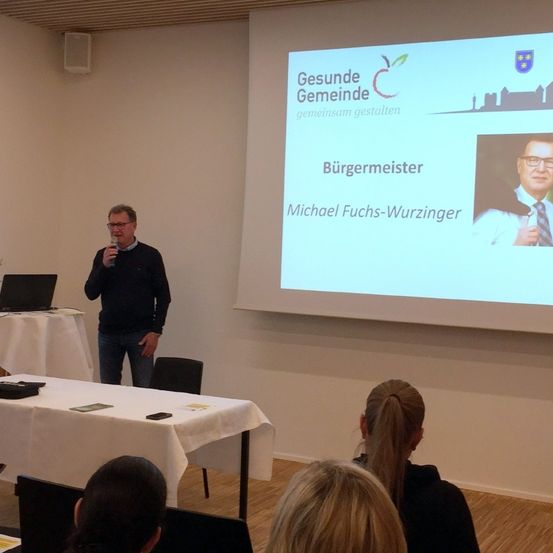 Ein Mann steht vor einer Projektionsleinwand, auf der eine Folie von Michael Fuchs-Wurzinger, dem Bürgermeister, zu sehen ist. Menschen sitzen um einen Tisch in einem Besprechungsraum.