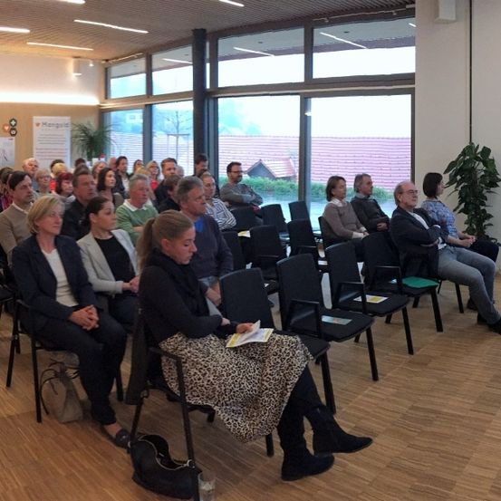Ein Seminarraum mit Holzböden und großen Fenstern, gefüllt mit Teilnehmern. Stühle sind in Reihen angeordnet, und die Teilnehmer sitzen, einige stehen. Eine Person liest von einem Stück Papier.