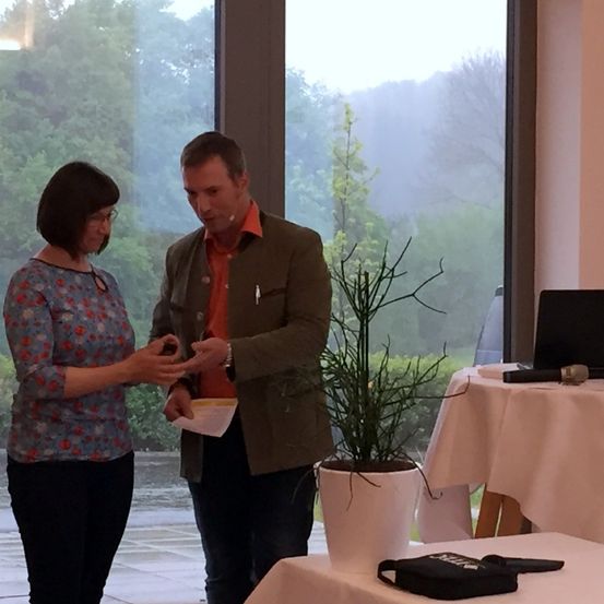 Ein Mann und eine Frau stehen vor einer Pflanze und halten Papiere. Sie sind drinnen mit großen Fenstern, die einen Garten zeigen.