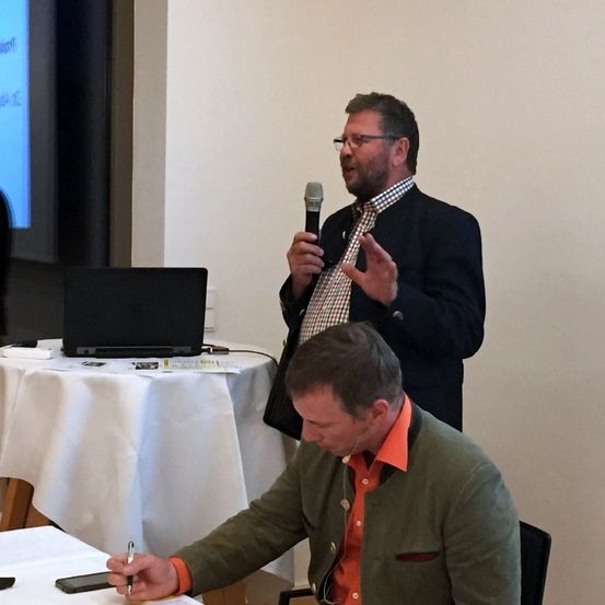 Ein Mann spricht in ein Mikrofon an einem Tisch, ein anderer Mann hört zu. Ein Laptop steht auf dem Tisch, und eine Projektionsleinwand befindet sich hinter dem Sprecher.