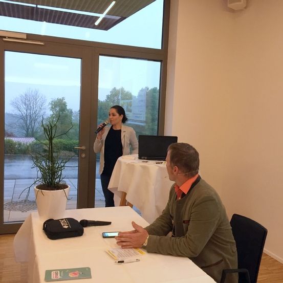 Ein Mann sitzt an einem Tisch und benutzt ein Telefon, während eine Frau hinter ihm in ein Mikrofon spricht. Ein Laptop und eine Pflanze im Topf stehen auf dem Tisch.