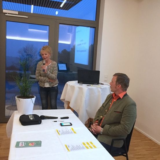 Eine Frau spricht in ein Mikrofon, während ein Mann an einem Tisch sitzt, beide sehen durch eine Glastür mit Blick auf Bäume. Ein Laptop steht auf einem Tisch hinter ihnen, und eine Projektionsleinwand ist sichtbar.