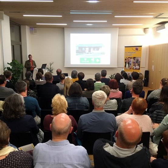 Ein Mann hält eine Präsentation in einem Raum mit einer Projektionsleinwand, während eine Menge von Menschen davor sitzt.