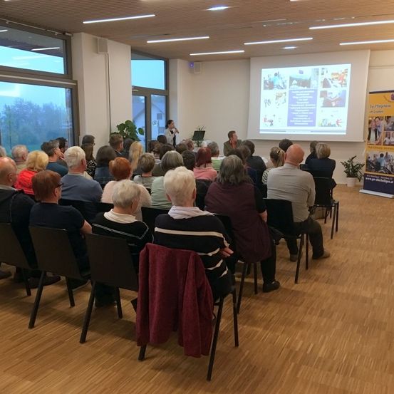 Eine Gruppe von Menschen sitzt in Stühlen um einen Raum herum und hört einer Präsentation zu. Eine Projektionsleinwand ist an der Wand sichtbar und zeigt eine Präsentation an.
