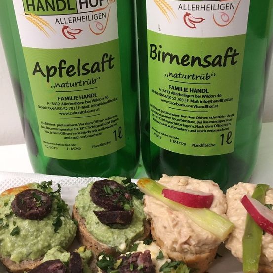 Zwei grüne Pflanzensäfte, beschriftet mit 'Apfelsaft naturtrüb' und 'Birnensaft naturtrüb', stehen nebeneinander. Darunter befindet sich ein Tablett mit verschiedenen kleinen Speisen, darunter Avocado, Radieschen und grüne Kräuter.