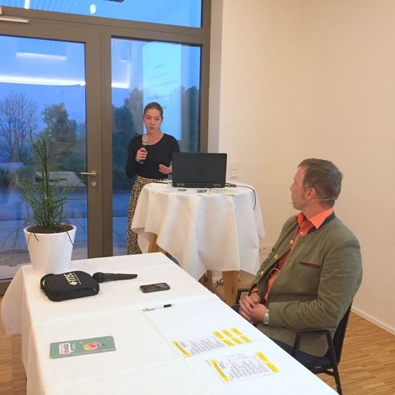 Eine Frau steht hinter einem Tisch mit einem Laptop und hält ein Mikrofon. Ein Mann sitzt vor ihr und hört zu. Ein Topf mit einer Pflanze steht links und Papiere liegen auf dem Tisch. Der Raum hat Glastüren und einen Holzboden.