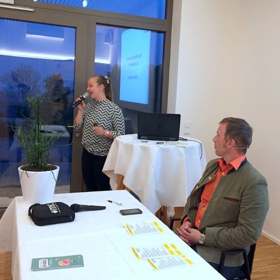 Eine Frau steht neben einem Mikrofon und spricht zu einem Publikum. Hinter ihr sitzt ein Mann an einem Tisch. Ein Laptop steht auf dem Tisch und eine Pflanze im Topf ist in der Nähe. Die Umgebung deutet auf eine Präsentation oder Besprechung hin.