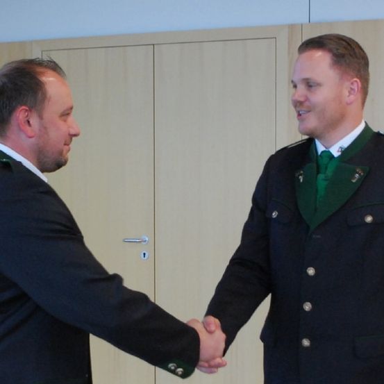 Zwei Männer, einer im Anzug und der andere in Uniform, schütteln sich die Hände in einem Raum mit hellen Holzverkleidungen.