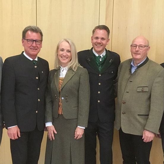 Fünf Personen stehen in einer Reihe, lächelnd, vor einer Holzwand. Sie tragen traditionelle deutsche Kleidung.