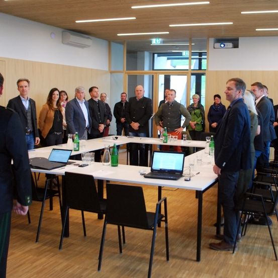 Eine Gruppe von Menschen steht um einen Konferenztisch mit Laptops, Getränken und Gläsern. Sie scheinen an einer Besprechung oder Diskussion teilzunehmen.