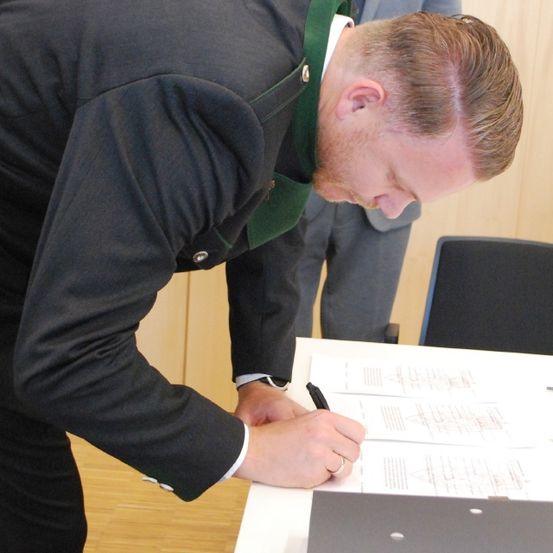 Ein Mann in Anzug unterzeichnet ein Dokument, während er über einem Schreibtisch gebeugt ist; ein Ordner und ein Stuhl sind in der Nähe.