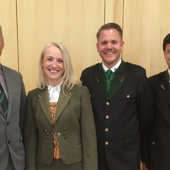 Vier Personen, zwei Männer und zwei Frauen, stehen nebeneinander vor einer Holzwand, alle in formeller Kleidung und lächelnd für ein Foto.
