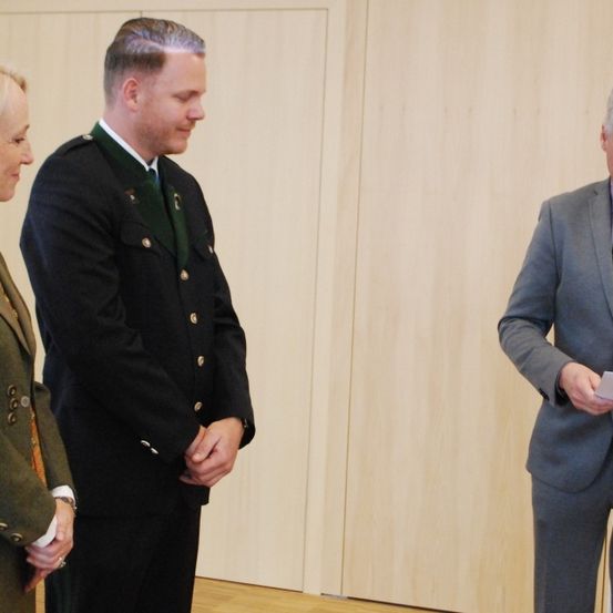 Drei Personen stehen in einem Raum; ein Mann in Uniform schaut einen Mann mit Papier an, während eine Frau in Uniform zuschaut.
