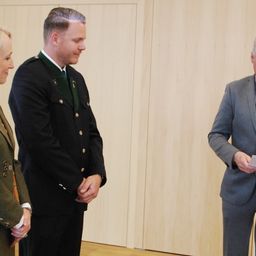 Drei Personen stehen in einem Raum; ein Mann in Uniform schaut einen Mann mit Papier an, während eine Frau in Uniform zuschaut.