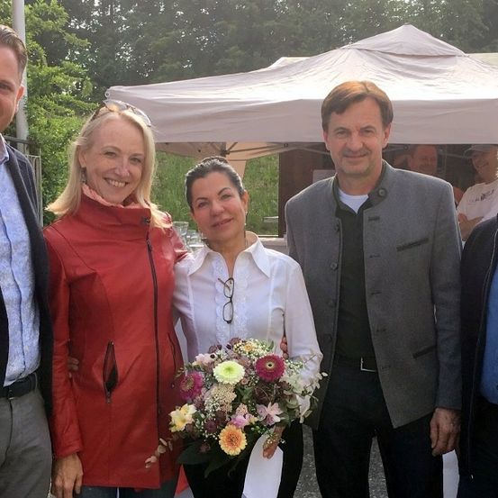 Fünf Menschen, die lächelnd für ein Foto posieren, mit einer Frau, die in der Mitte einen Blumenstrauß hält. Hinter ihnen ist ein weißes Zelt und Bäume zu sehen.