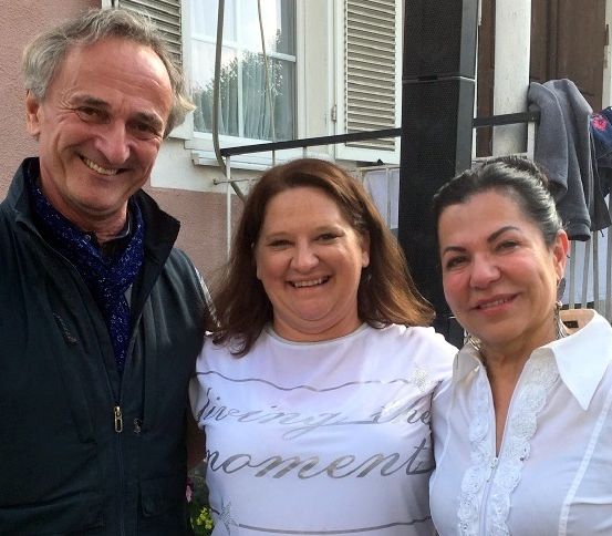 Drei Menschen lächeln vor einem Gebäude, wobei eine Frau ein weißes Shirt mit 'Genieße den Moment' trägt und ein Mann einen schwarzen Mantel.