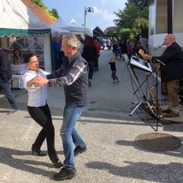 Ein Paar tanzt im Freien, während ein Musiker in der Nähe ein Instrument spielt. Es gibt Zelte und Leute in der Umgebung.