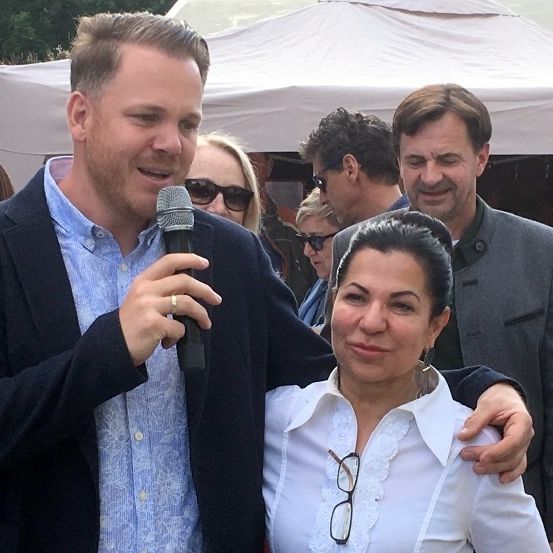 Ein Mann mit einem Mikrofon steht neben einer Frau bei einer Veranstaltung im Freien, mit einem weißen Zelt im Hintergrund und mehreren Personen in der Nähe.