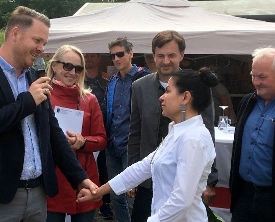 Mehrere Menschen stehen unter einem weißen Zelt. Ein Mann hält ein Mikrofon, während eine Frau ein Papier in der Hand hat. Ein anderer Mann und eine Frau halten Händchen, während ein Mann neben ihnen lächelt.
