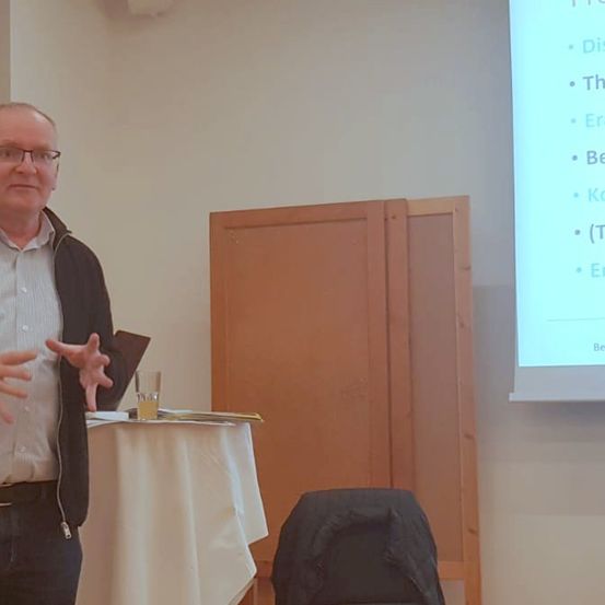 Ein Mann steht vor einem Schreibtisch, möglicherweise in einem Klassenzimmer. Er spricht und macht Handbewegungen. Hinter ihm befindet sich eine weiße Wand mit einem Holzschrank und einem Projektionsbildschirm.