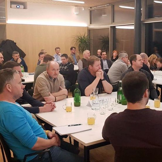 Eine Gruppe von Männern sitzt in einem Konferenzraum um einen Tisch herum. Der Tisch ist mit Getränken und einem Notizblock gefüllt. Eine Projektionslampe ist an der Decke zu sehen.