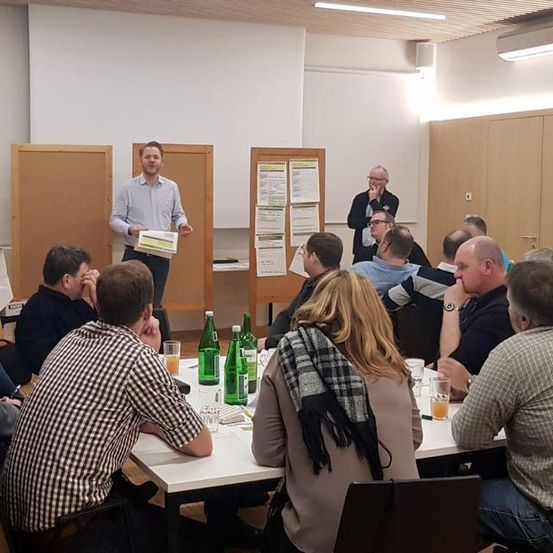 Eine Gruppe von Menschen sitzt um einen Tisch in einem Raum. Ein Mann steht vor ihnen und hält ein Papier. Auf dem Tisch stehen Flaschen und Gläser. Einige Menschen tragen Brillen und Uhren.